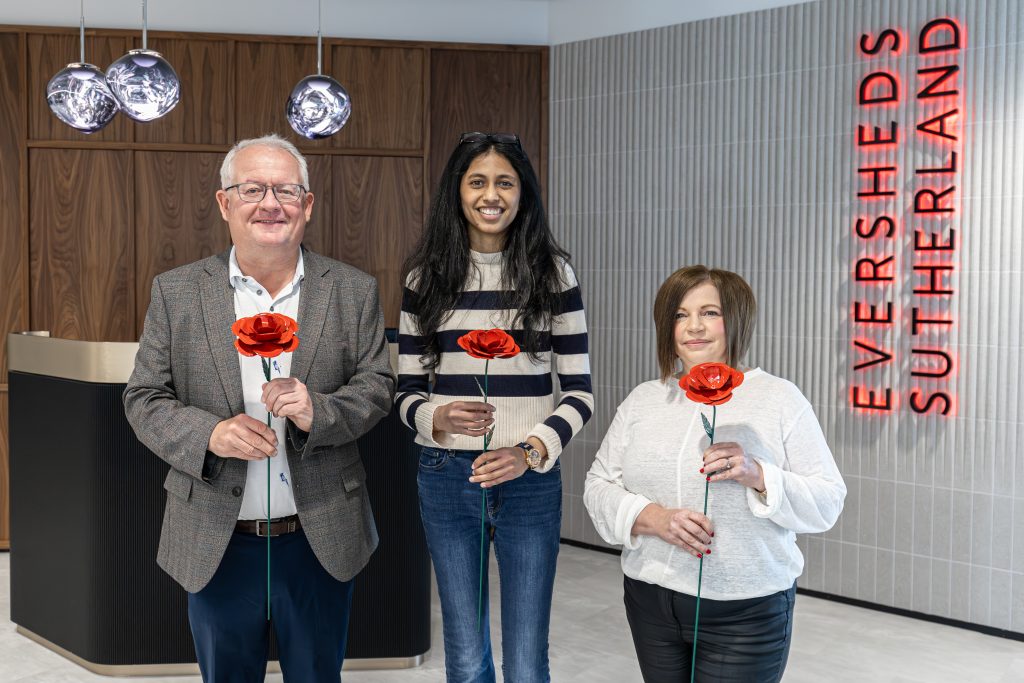Three people hold metal red roses in the reception area of an office building.