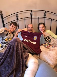 A man holding up an Aston Villa shirt as he sits in bed with his children either side of him