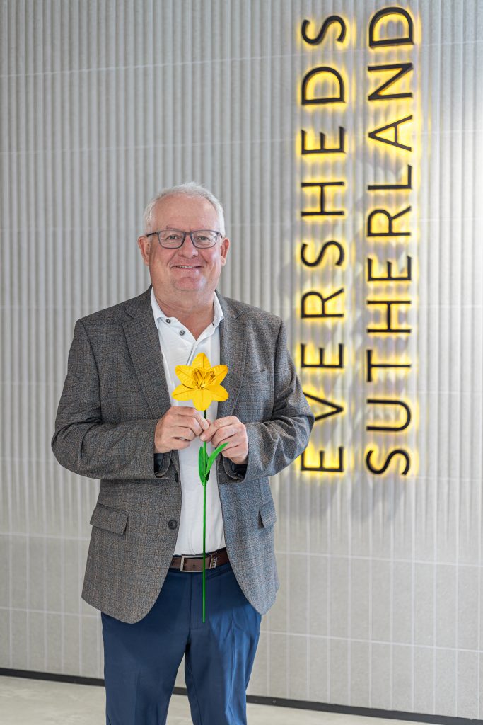 A man in a suit holds a daffodil in front of a yellow Eversheds Sutherland sign