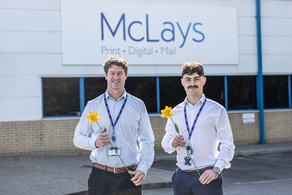 Two men wearing lanyards hold daffodils as they stand outside McLays