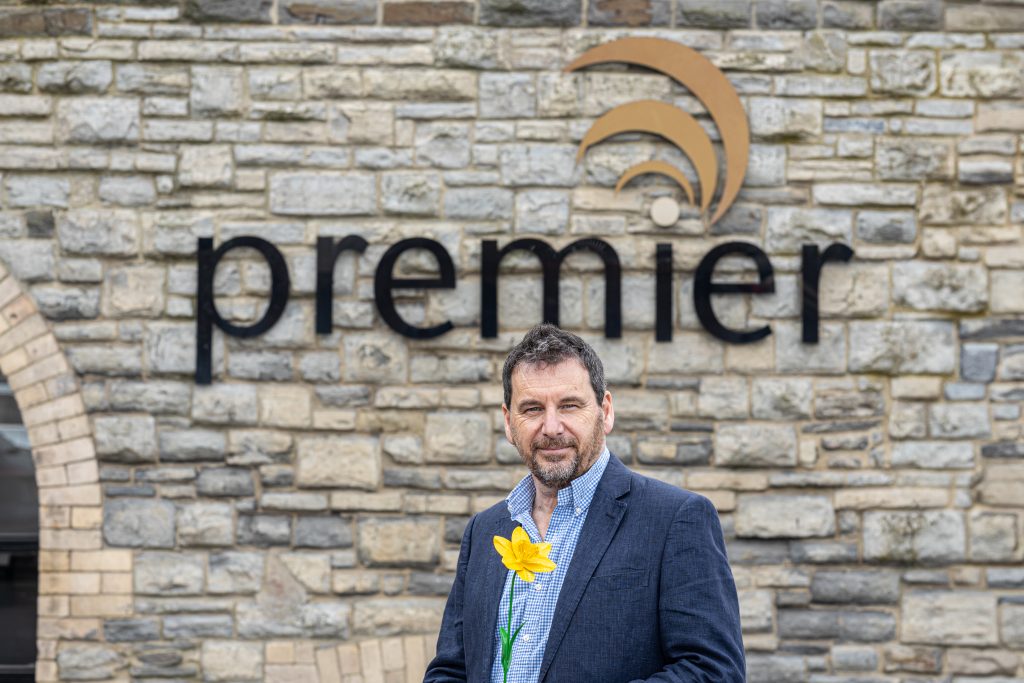A bearded man holds a daffodil in front of a brick wall with the Premier logo on it.