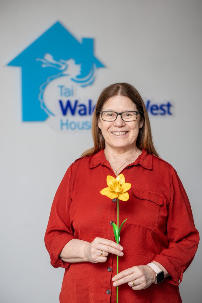 A woman in a red top holds a daffodil as she stands in front of a bilingual Wales & West Housing sign